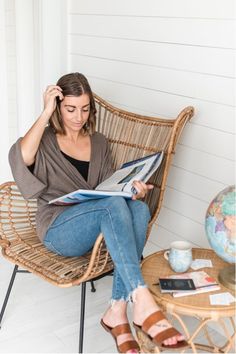 a woman sitting in a wicker chair reading a book while holding a coffee cup