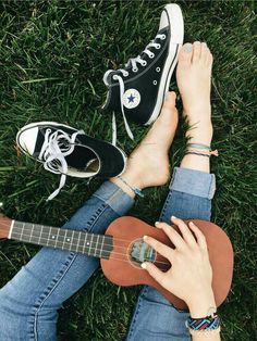 a person sitting in the grass with their feet on an ukulele and wearing converse shoes