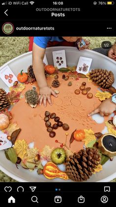 a child's hands are on the top of a table covered in autumn leaves and pine cones