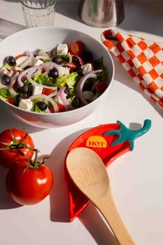 a salad in a bowl next to a wooden spoon on a white table with a checkered napkin