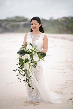 a woman standing on top of a sandy beach holding a bouquet of flowers and greenery