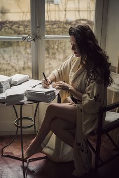 a woman sitting at a table writing on a piece of paper next to a window