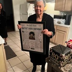 an older woman holding up a newspaper in her kitchen while standing next to a cake