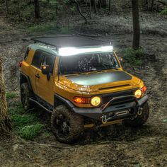 a yellow jeep is parked in the mud near a tree and some trees with its lights on