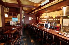 a woman standing at the bar in a restaurant with lots of wooden tables and chairs