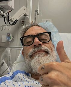 a man with glasses and a beard in a hospital bed giving the thumbs up sign