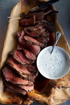 a wooden platter filled with sliced meat and dipping sauce next to a bowl of ranch dressing