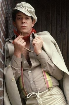 a man in a suit and hat is holding something up to his ear while leaning against a wall