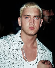 a young man with blonde hair wearing a white shirt and silver necklace, looking at the camera