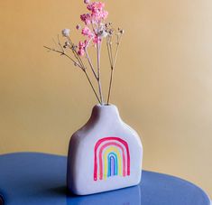 a white vase with pink flowers and a rainbow painted on the side sitting on a blue table