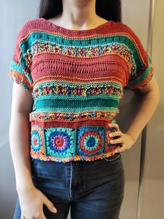 a woman wearing a colorful crochet top with her hands on her hips, standing in front of a wall