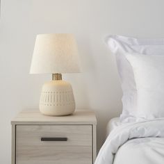 a nightstand with a lamp on top of it next to a white bed and pillows