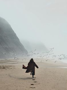 a woman walking on the beach with birds flying around her