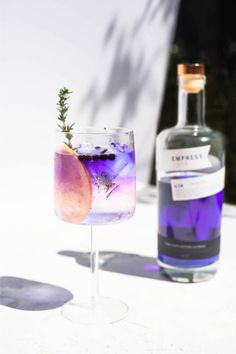 a purple drink sitting next to a bottle on top of a white tablecloth with a sprig of rosemary in it