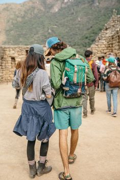 two people with backpacks walking in front of some stone structures and mountains, one is carrying a back pack