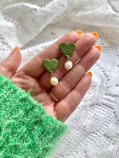 a hand holding two heart shaped earrings on it's fingers with green and white accents