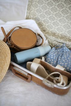 an open suitcase filled with clothes and other items on top of a white bed next to a straw hat