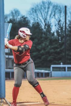 a woman holding a baseball bat while standing on top of a dirt field with trees in the background