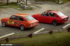 two red cars parked next to each other on a dirt road in front of a wooden fence