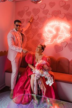 a man and woman dressed in pink posing for the camera with hearts on the wall behind them