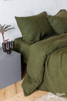 a bed with green sheets and pillows next to a small plant on a wooden floor