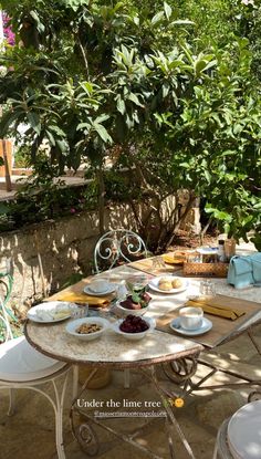 an outdoor table with plates and bowls of food on it in the shade of some trees