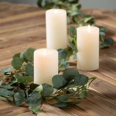 three white candles sitting on top of a wooden table next to eucalyptus leaves and greenery