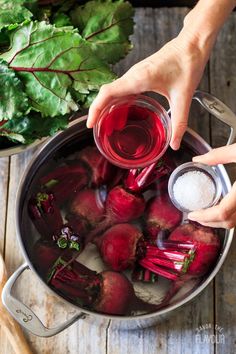 a person pouring red liquid into a pot filled with beets and greens on top of a wooden table