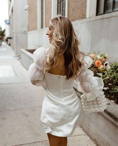 a woman is walking down the street with flowers in her hand and wearing a white dress