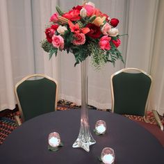 a tall vase filled with red and pink flowers on top of a table next to two green chairs