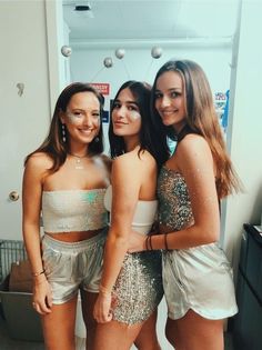 three young women standing next to each other in front of a mirror wearing silver outfits