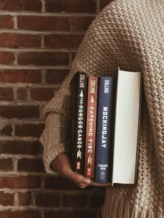 a person is holding three books in front of a brick wall and wearing a sweater