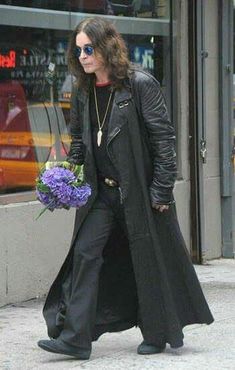 a man walking down the street with flowers in his hand and wearing a long black coat