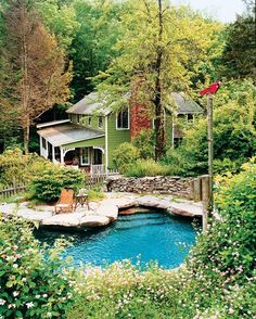 a house in the woods next to a pond with chairs on it and trees around