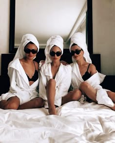 three women in white robes sitting on a bed wearing sunglasses and towel wrapped around their heads