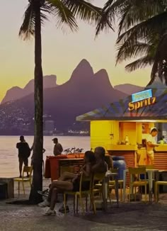 people sitting at tables near the water with mountains in the background and palm trees around them