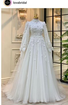 a white wedding dress with long sleeves on display in front of a mirror and plant