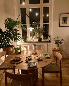 a dining room table with plates and cups on it in front of a window filled with potted plants