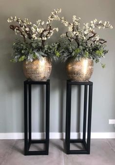 two metal vases with flowers in them sitting on top of black stands against a gray wall