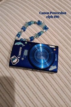 a blue camera sitting on top of a bed next to a beaded necklace with charms