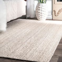 a large white rug in the middle of a room next to a bed and vase with flowers