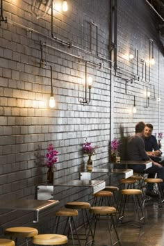 people sitting at tables in a restaurant with lights on the brick wall and wooden stools