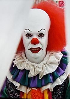 a creepy clown with red hair and white face is standing in front of the camera