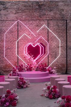 a heart - shaped neon sign surrounded by flowers and stools in front of a brick wall