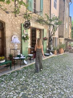 a woman walking down a cobblestone street in front of a building with green shutters