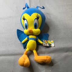 a blue and yellow bird stuffed animal laying on top of a gray bed sheet with a tag in it's ear