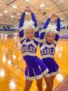 two cheerleaders in blue and white outfits standing on a basketball court with their hands up