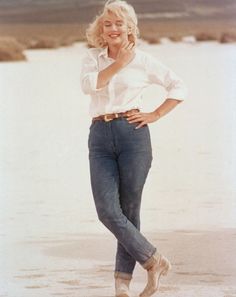 a woman standing on the beach with her hands in her pockets and smiling at the camera