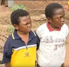 two young boys standing next to each other on a dirt ground with grass in the background