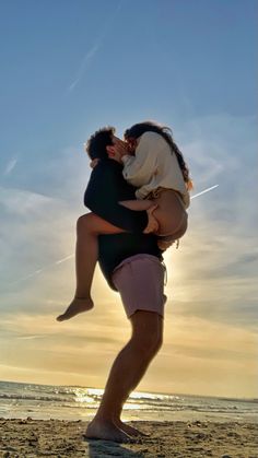 a man and woman hug on the beach as the sun sets in the sky behind them